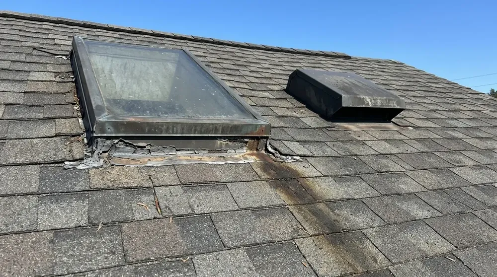 Skylight with cracked, peeling flashing and a water stain running down the shingles next to a roof vent